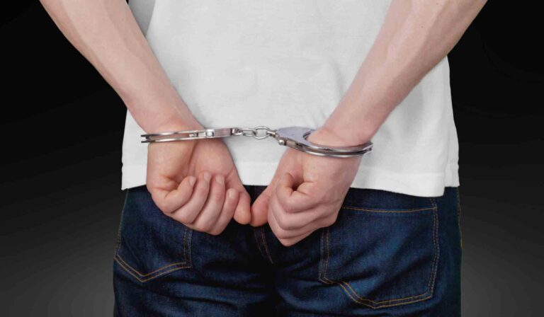Person in handcuffs with hands behind their back, wearing a white shirt and jeans.
