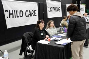 A student speaks with volunteers seated at tables during a financial literacy event, with “Child Care” and “Clothing” station signs displayed behind them.