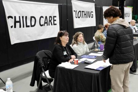 A student speaks with volunteers seated at tables during a financial literacy event, with “Child Care” and “Clothing” station signs displayed behind them.