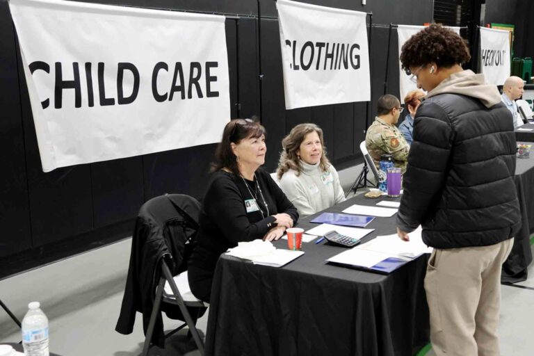 A student speaks with volunteers seated at tables during a financial literacy event, with “Child Care” and “Clothing” station signs displayed behind them.