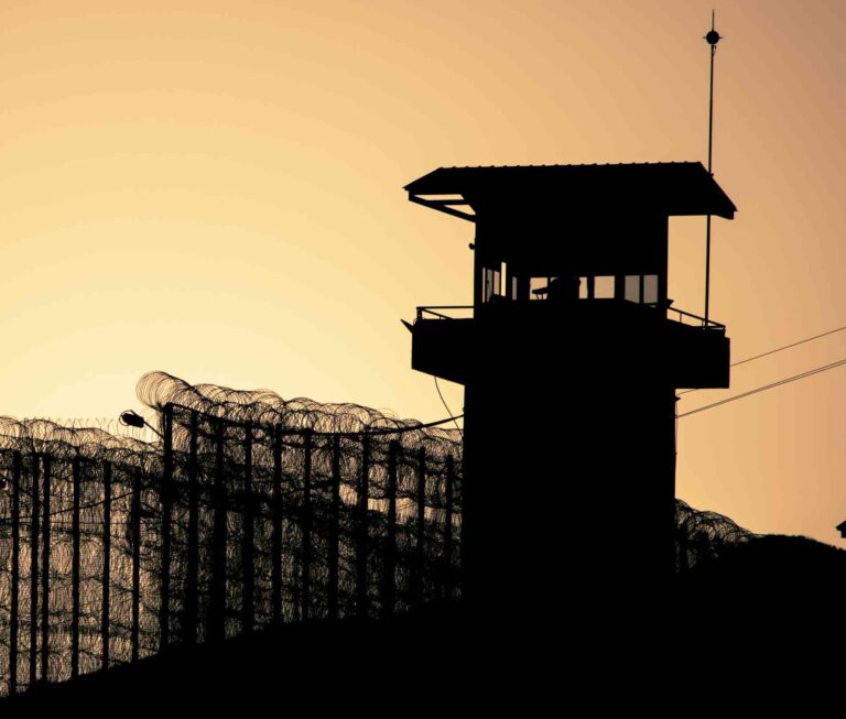 Silhouette of a prison watch tower and perimeter fencing at sunset.