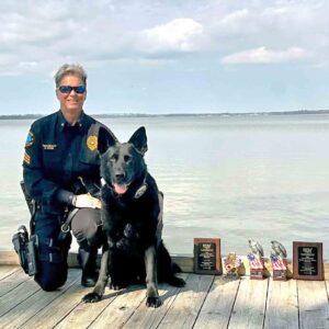 Sergeant Kristin Pond kneels beside K-9 Kodi on a waterfront dock with competition awards displayed in front of them.