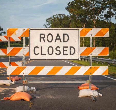 Road closed sign mounted on orange and white barricades blocking a roadway