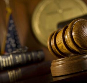 Wooden judge’s gavel resting on a sound block with law books, a clock, and an American flag in the background.