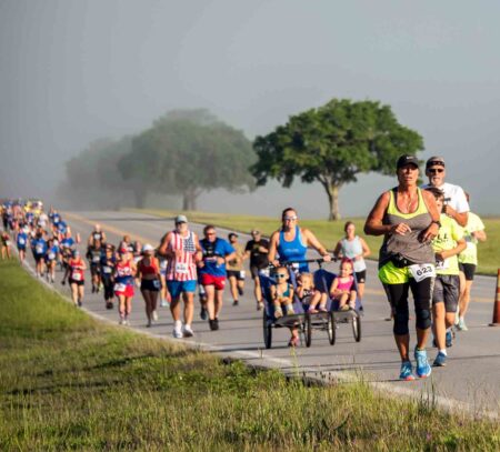 Participants run and walk along a roadway during the Eglin Gate-to-Gate Memorial Run/Walk, with multiple runners and walkers moving in a group across the base.
