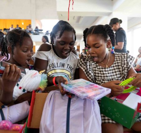 Three girls open gift-filled shoeboxes and smile as they look at toys and school supplies inside.