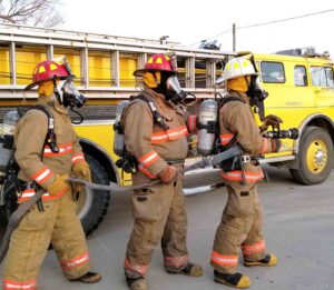 Firefighters wearing full protective gear and breathing apparatus operate a hose line beside a yellow fire engine during a training exercise.