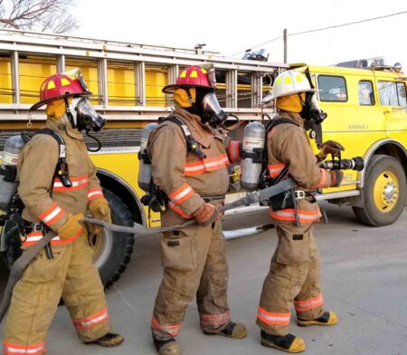 $5,000 scholarship offered for future firefighters, EMTs Firefighters wearing full protective gear and breathing apparatus operate a hose line beside a yellow fire engine during a training exercise.