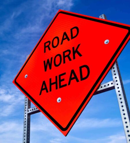 Orange “Road Work Ahead” warning sign mounted on a roadside post against a blue sky.