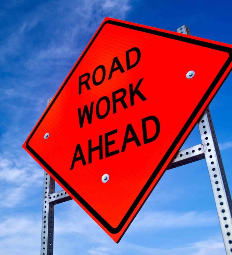 Orange “Road Work Ahead” warning sign mounted on a roadside post against a blue sky.
