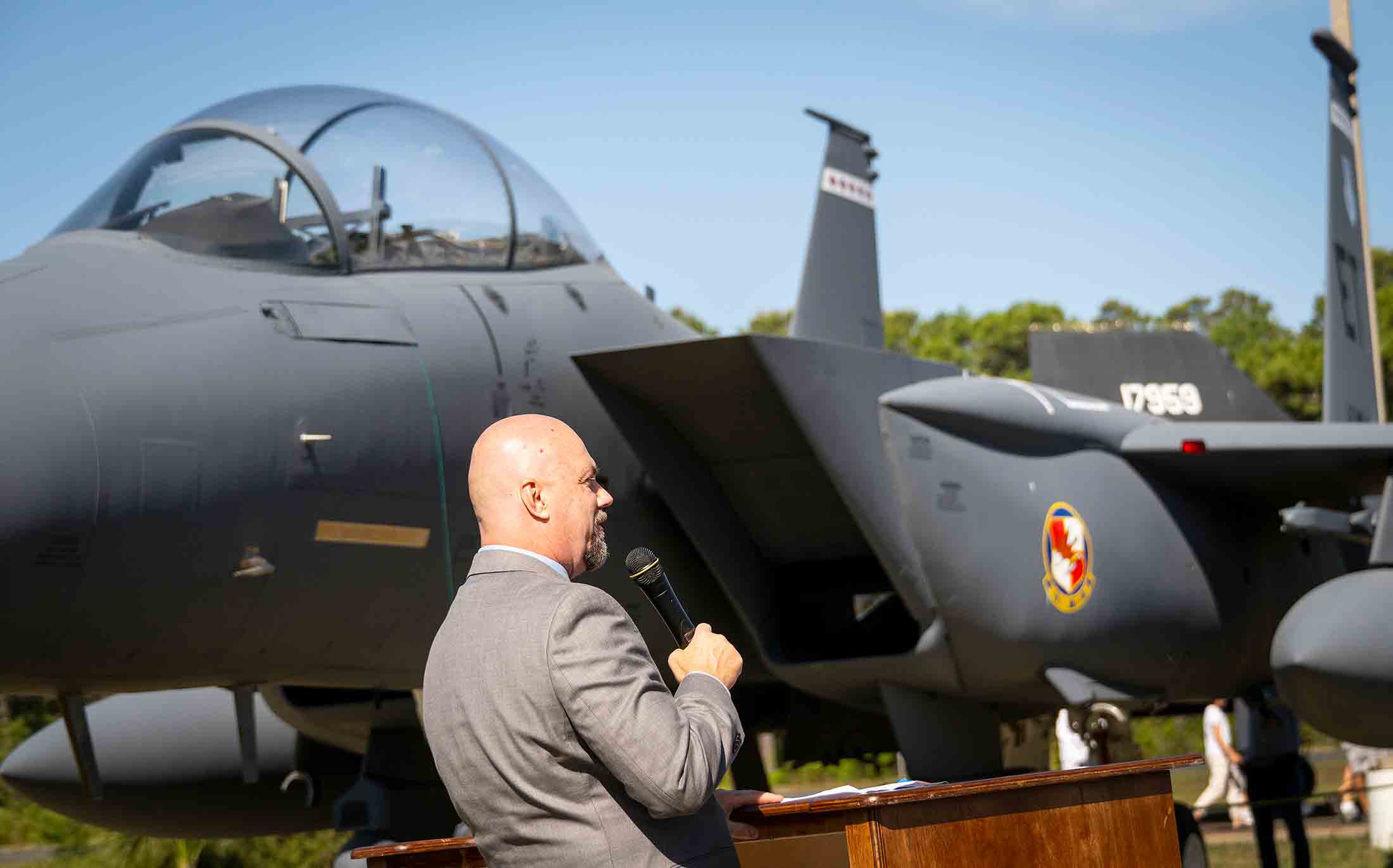 Museum director speaks at podium beside an F-15E Strike Eagle during a dedication ceremony