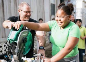Elementary student uses a miter saw with guidance from an instructor during a hands-on math activity