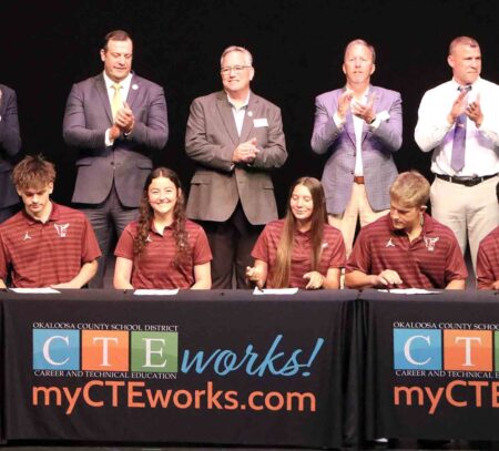 Okaloosa County students sign commitment letters during the 2026 CTE Signing Day ceremony as school officials stand behind them and applaud