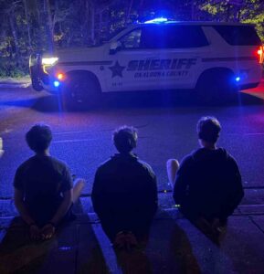 Three juveniles sit handcuffed on a curb at night as Okaloosa County Sheriff’s deputies stand nearby with a patrol vehicle and flashing lights.