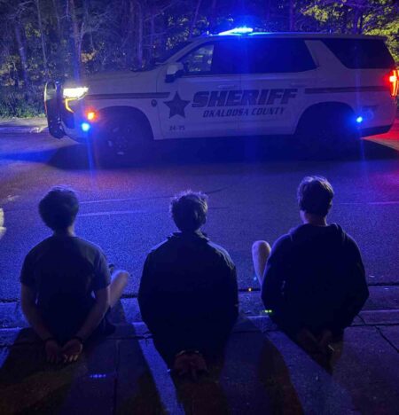 Three juveniles sit handcuffed on a curb at night as Okaloosa County Sheriff’s deputies stand nearby with a patrol vehicle and flashing lights.