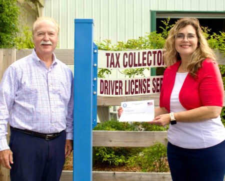 Okaloosa County Tax Collector Ben Anderson and customer service agent Hiedi Jacobs stand outside a tax collector office location while displaying a letter of recognition from a U.S. Air Force lieutenant colonel praising Jacobs for assisting military families with vehicle registration and title transfers.