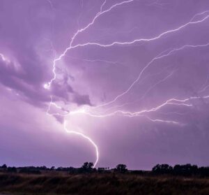 A bright cloud-to-ground lightning strike branches across a stormy purple sky above a rural landscape at night.