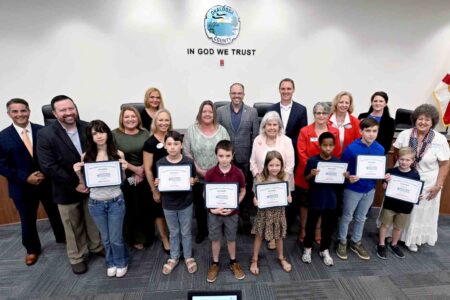Recipients of the 2026 Okaloosa Saves Teach a Child to Save campaign pose with Okaloosa County commissioners, Okaloosa Saves board members and participating financial institution representatives after being recognized during the July 21, 2026, Okaloosa County Commission meeting in Fort Walton Beach.