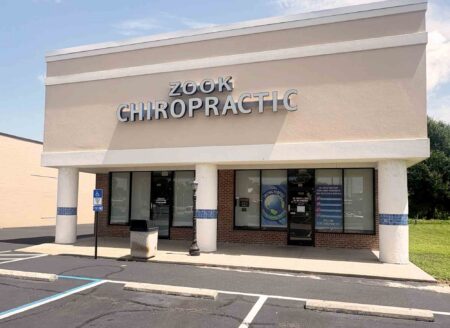 Exterior front entrance of Zook Chiropractic clinic in a beige retail building with large "Zook Chiropractic" signage above the storefront, covered walkway, and parking spaces in front.