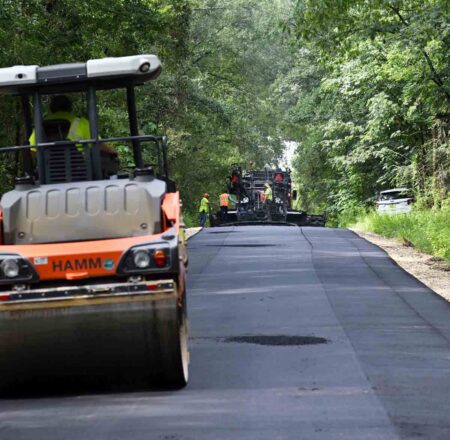Infrastructure surtax helps pave 180 miles of Okaloosa roads Road crews pave a rural Okaloosa County road as a roller follows behind the paving equipment.