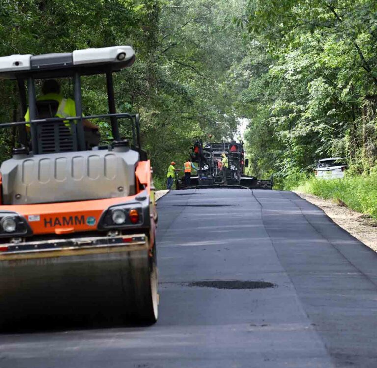 Road crews pave a rural Okaloosa County road as a roller follows behind the paving equipment.